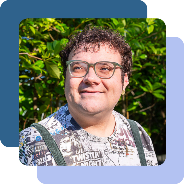 Connor is a young white non-binary person with short brown curly hair and light facial hair. They are pictured smiling wearing green glasses and green dungarees over a newsprint pattern t-shirt.