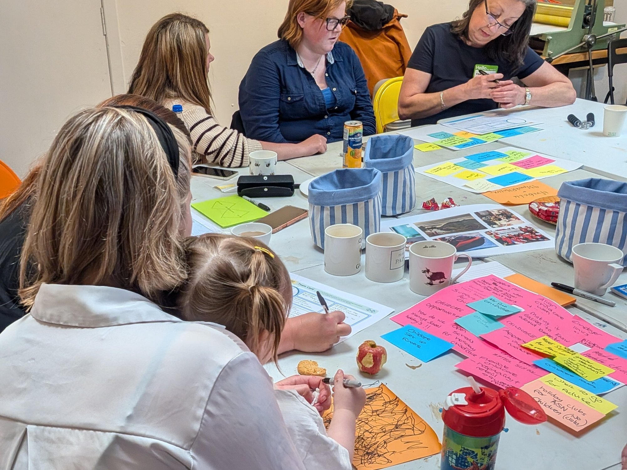Looking over the shoulder of a Core Skills for Employability learner in Stranraer sitting at a table with her daughter on her lap, pictured doodling on a large sticky note next to half-eaten apple. The table is filled with mugs, paper, and coloured sticky notes and surrounded by other learners. Looking over the shoulder of a Core Skills for Employability learner in Stranraer sitting at a table with her daughter on her lap, pictured doodling on a large sticky note next to half-eaten apple. The table is filled with mugs, paper, and coloured sticky notes and surrounded by other learners.