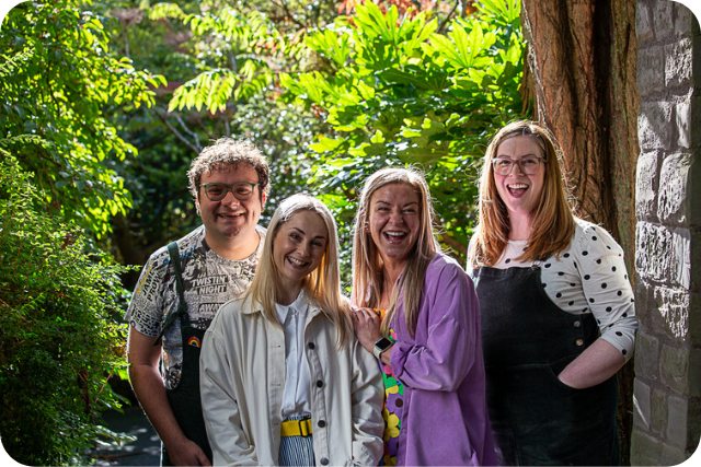 Connor, Jo, Katie, and Kim smiling together in front of sunny greenery.