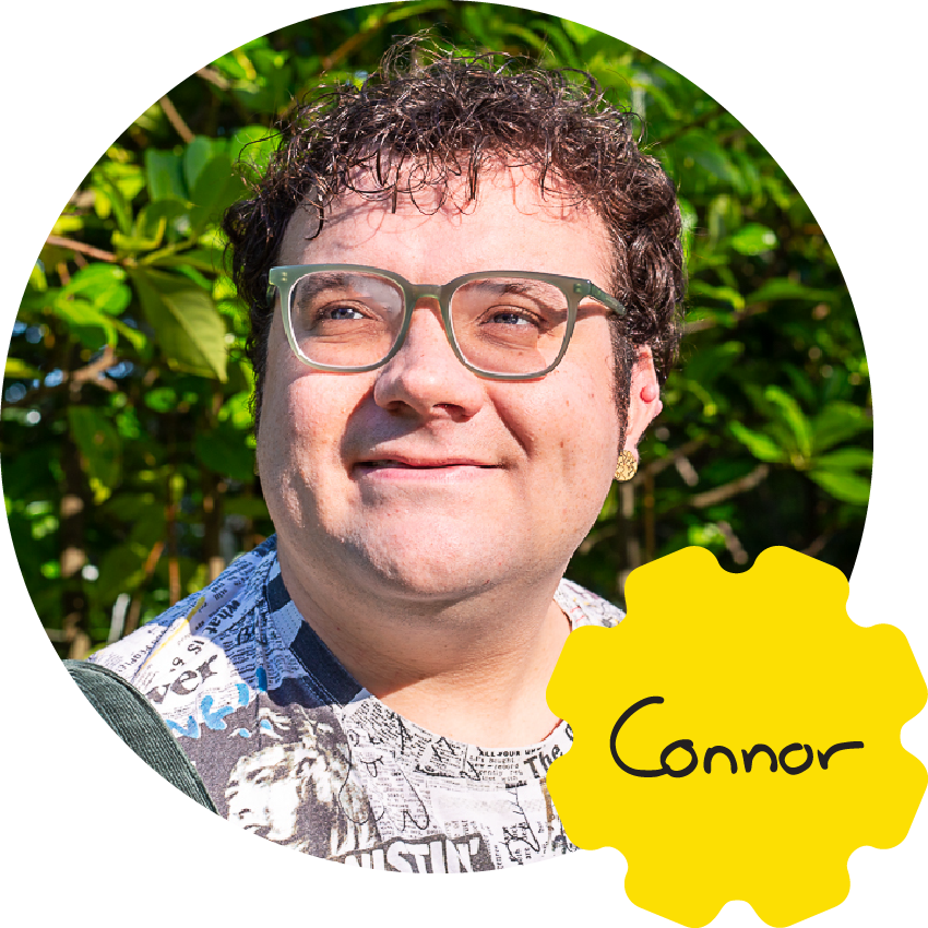 Connor is a young white non-binary person with short brown curly hair and light facial hair. They are pictured smiling wearing green glasses and green dungarees over a newsprint pattern t-shirt. Their name is handwritten in the corner with rounded letters and the capital C joined to the small O.