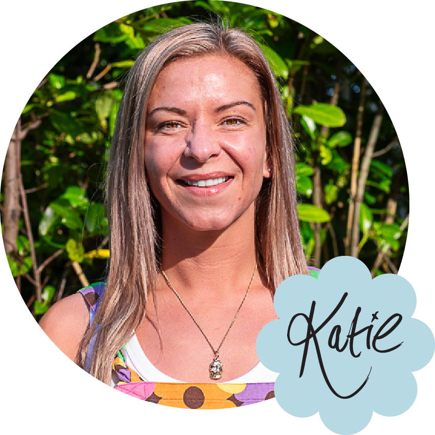 Katie is a young white woman with straight blonde hair. She's pictured here smiling wearing multi-coloured floral dungarees. Her name is handwritten in the corner with a elaborate curly K with a swooping curve in the kick, joined up curvy small letters, and with an x dotting the i.