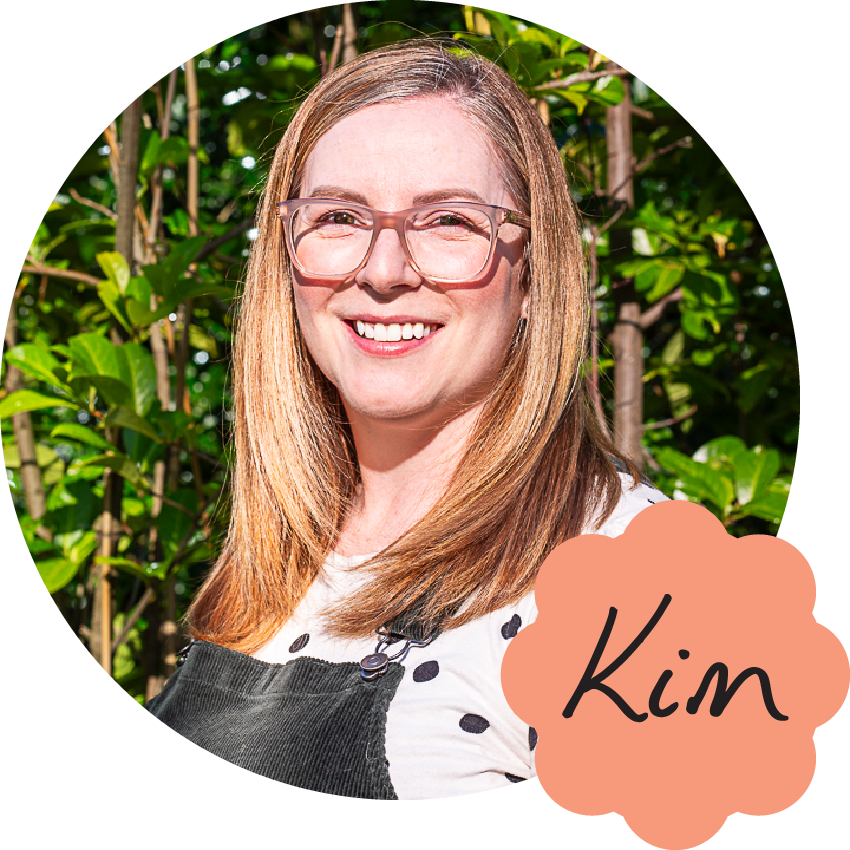 Kim is a middle-aged white woman with wavy brown hair. She's pictured here smiling, wearing glasses, lip gloss and her signature black pinafore dress over a white polka dot blouse. Her name is handwritten in the corner with a tall K with a curve in the kick, and flicks on the tails of the i and m.