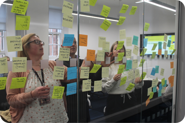 A view through glass and coloured sticky notes showing people looking at and adding notes on the other side during a workshop.