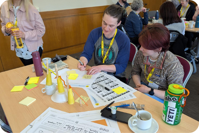 Staff from Angus Alive writing intently while sitting around a table filled with models, paper templates, sticky notes, and various refreshments.